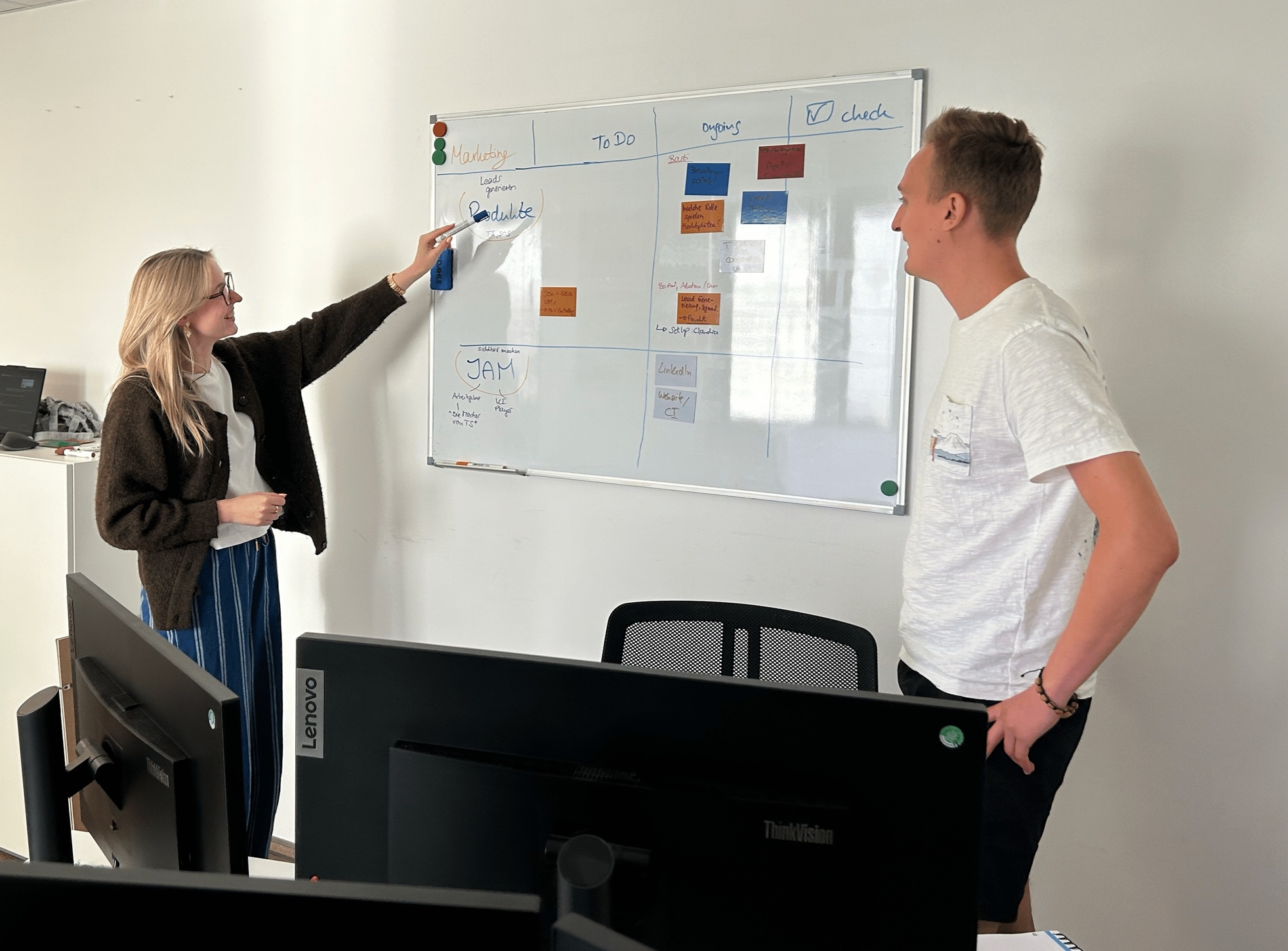 Two colleagues discuss tasks at a whiteboard in an office.