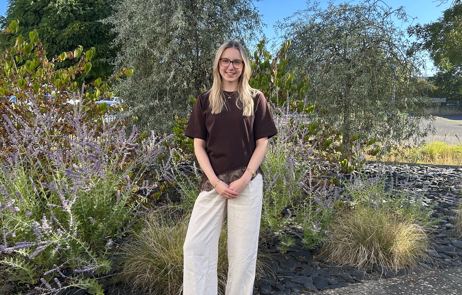 A blonde woman stands in a garden, smiling at the camera in front of green plants.