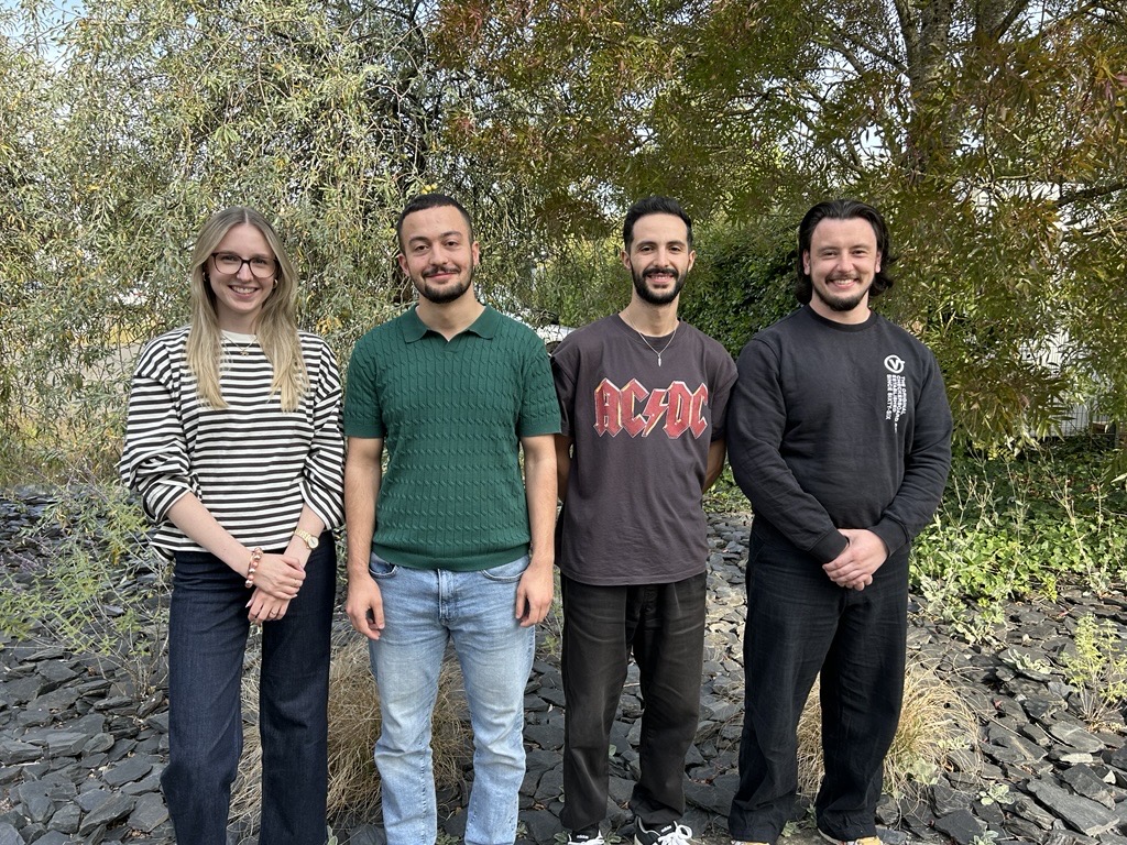 Four young people are standing in a garden and smiling at the camera. There is one woman and three men.